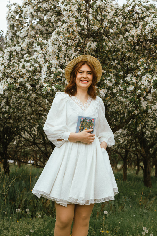 Woman in the Avonlea Dress standing among flowering trees, holding a book and wearing a straw hat.