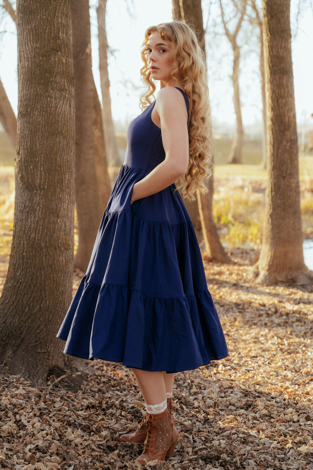 Woman in a blue dress standing in a forest with trees and ground covered in leaves.