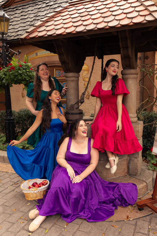 Four women in Gemstone Dress Collection posing in Sapphire Blue, Amethyst Purple, Ruby Red, and Tourmaline Teal in a storybook village